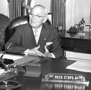 Black and white photo of Harry S. truman in the Oval Office, with his desk sign reading "The Buck Stops Here!" prominently shown.